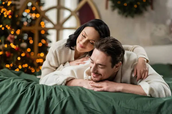Retrato de casal alegre com olhos fechados deitados juntos na cama perto da árvore de Natal — Fotografia de Stock