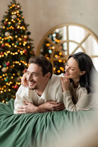 Портрет мечтательной пары, смотрящей в сторону и лежащей вместе на кровати возле рождественской елки с огнями — стоковое фото