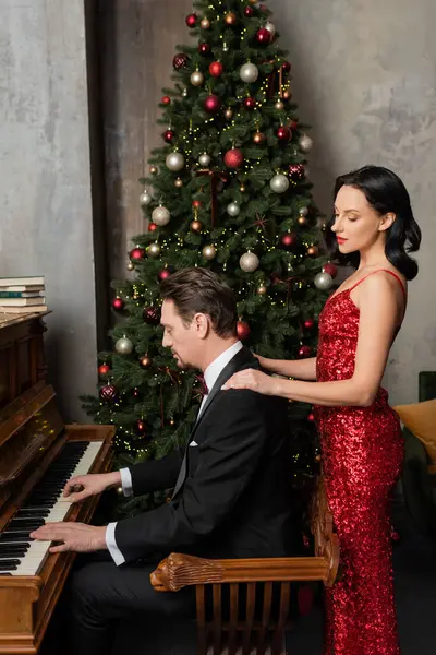 Pareja rica, mujer morena en vestido rojo de pie cerca de marido tocando el piano, Feliz Navidad - foto de stock