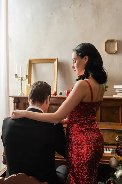 Багата пара, приваблива жінка в червоній сукні, що стоїть поруч з чоловіком в тукседо грає на піаніно — стокове фото