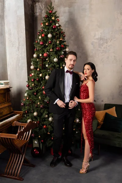 Familia rica, mujer elegante en vestido rojo de pie cerca del hombre en esmoquin, piano y árbol de Navidad - foto de stock