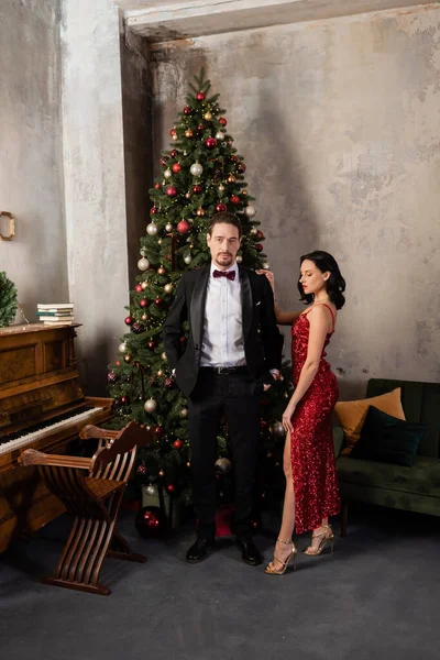 Pareja rica, mujer elegante en vestido rojo de pie cerca del hombre en esmoquin, piano y árbol de Navidad - foto de stock