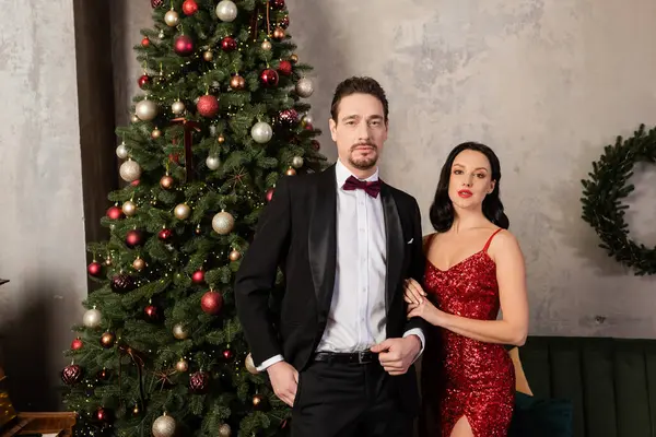 Pareja rica, mujer elegante en vestido rojo de pie cerca del hombre en esmoquin y árbol de Navidad decorado - foto de stock