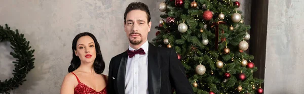 Couple riche, femme élégante en robe rouge debout près de l'homme en smoking et arbre de Noël, bannière — Photo de stock