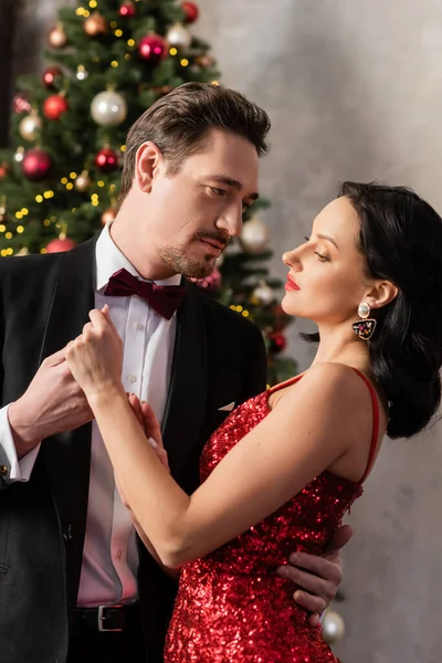 Hombre guapo en traje de la mano de la mujer bonita en vestido elegante rojo cerca del árbol de Navidad - foto de stock
