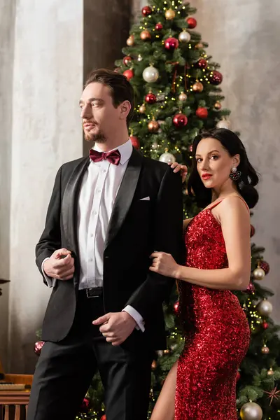 Bonita mujer con pelo morena de pie en vestido rojo cerca de hombre guapo en traje y árbol de Navidad - foto de stock