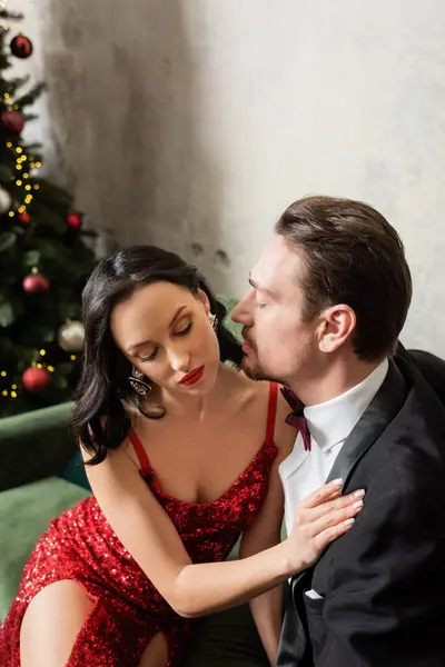 Gentleman en smoking assis sur canapé et embrasser la joue de la femme en robe rouge près de l'arbre de Noël — Photo de stock
