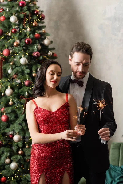 Beautiful wealthy couple in formal and elegant attire holding bright sparklers near Christmas tree — Stock Photo
