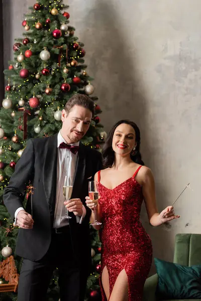 Rico casal feliz em traje formal segurando copos de champanhe e brilhos perto da árvore de Natal — Fotografia de Stock
