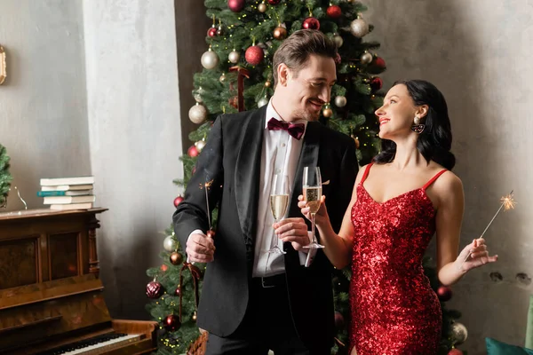 Riche couple joyeux en tenue formelle tenant des verres de champagne et des étincelles près de l'arbre de Noël — Photo de stock