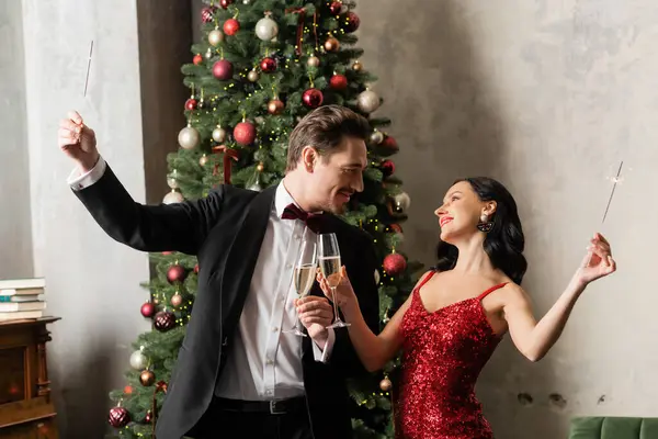 Joyful married couple in formal attire holding champagne glasses and sparklers near Christmas tree — Stock Photo