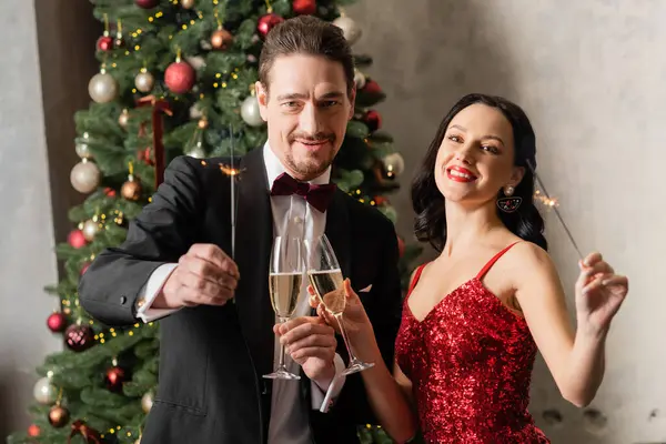 Heureux couple marié en tenue formelle tenant des verres de champagne et des étincelles près de l'arbre de Noël — Photo de stock