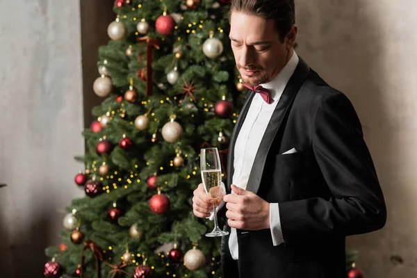 Homem rico bonito em smoking com gravata borboleta segurando copo de champanhe perto da árvore de Natal decorada — Fotografia de Stock