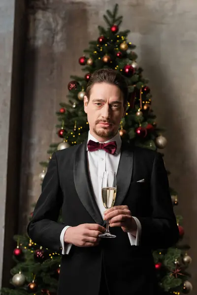 Cavalheiro rico em terno com gravata borboleta segurando copo de champanhe perto da árvore de Natal decorada — Fotografia de Stock