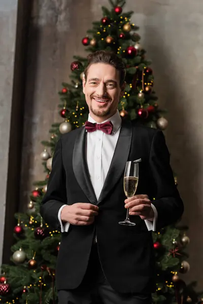 Cavalheiro alegre em terno com laço segurando copo de champanhe perto da árvore de Natal decorada — Fotografia de Stock