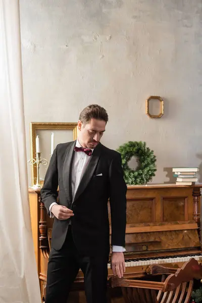 Cavalheiro rico em traje formal com gravata borboleta em pé perto de piano e coroa de Natal na parede — Fotografia de Stock