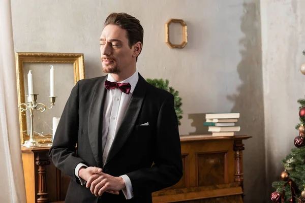 Hombre rico en traje formal con pajarita de pie cerca del piano en la víspera de Navidad, vacaciones de invierno - foto de stock