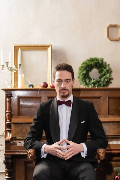 Elegant gentleman in formal attire with bow tie sitting near piano on Christmas eve, holiday — Stock Photo