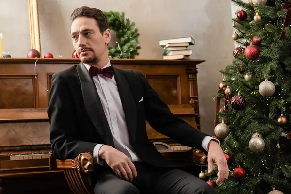 Елегантний джентльмен в офіційному вбранні з краваткою з бантом, що сидить біля фортепіано і прикрашена ялинка — стокове фото