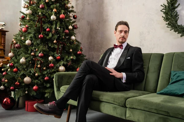 Well-dressed wealthy man with beard wearing tuxedo with bow tie sitting on sofa near Christmas tree — Stock Photo