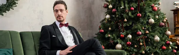 Uomo ben vestito con barba che indossa smoking con papillon seduto sul divano vicino all'albero di Natale, striscione — Foto stock