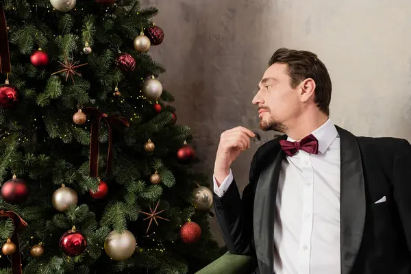 Caballero bien vestido con barba usando esmoquin con pajarita sentado en el sofá cerca del árbol de Navidad - foto de stock