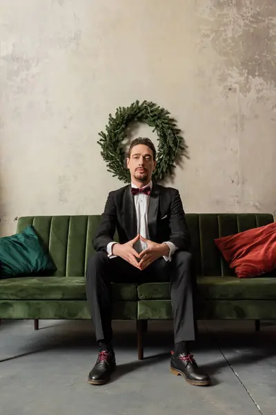 Gut gekleideter Herr mit Bart im Smoking mit Fliege sitzt auf Sofa in der Nähe des Weihnachtskranzes — Stockfoto