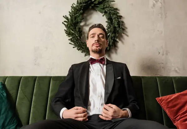 Riche gentleman avec barbe portant smoking avec noeud papillon assis sur canapé près de couronne de Noël — Photo de stock