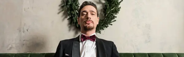 Homme riche avec barbe portant smoking avec noeud papillon regardant la caméra près de couronne de Noël, bannière — Photo de stock