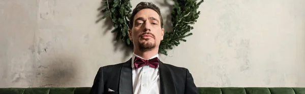Uomo ricco con barba indossa smoking con papillon guardando la macchina fotografica vicino alla corona di Natale, striscione — Foto stock