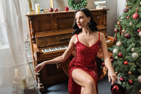 Schöne Frau in elegantem roten Kleid neben Klavier und geschmücktem Weihnachtsbaum sitzend, wohlhabendes Leben — Stockfoto