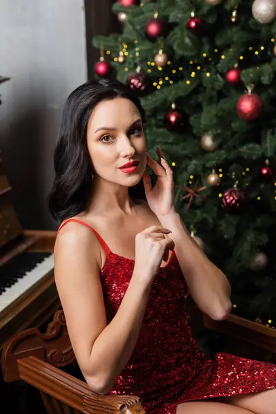 Mulher sofisticada em vestido vermelho sentado perto de piano e árvore de Natal decorada, vida rica — Fotografia de Stock