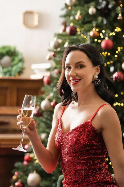 Веселая женщина в красном платье с бокалом шампанского рядом с украшенной елкой, богатая жизнь — стоковое фото