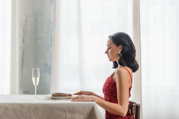 Vista lateral de la mujer elegante en vestido rojo sentado en la mesa de comedor con copa de champán y filete - foto de stock