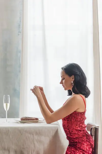 Vista lateral da mulher bonita em vestido vermelho sentado na mesa de jantar com copo de champanhe e bife — Fotografia de Stock