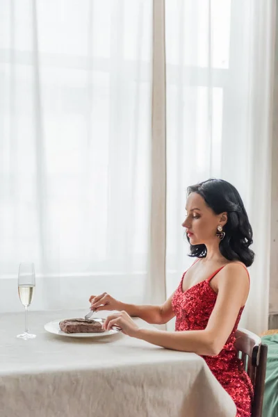 Elegant woman in red dress sitting at dining table with glass of champagne and cutting beef steak — Stock Photo