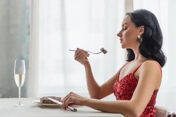 Elegant woman in red dress sitting at dining table with glass of champagne and eating beef steak — Stock Photo