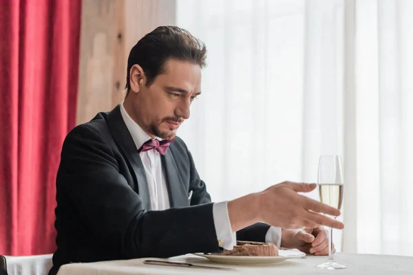 Красивый мужчина в смокинге сидит за обеденным столом с бокалом шампанского и бифштексом на тарелке — стоковое фото