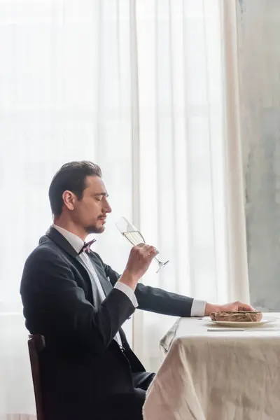 Homem bonito em smoking apreciando o sabor de champanhe e sentado à mesa com bife de vaca no prato — Fotografia de Stock