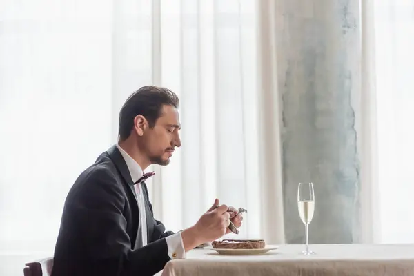 Hombre guapo en esmoquin disfrutando del sabor de carne de res en el plato cerca de champán en vidrio en la mesa de comedor - foto de stock