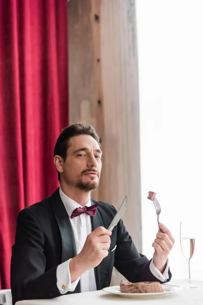 Ricco gentiluomo in smoking godendo il gusto della bistecca di manzo vicino champagne in vetro sul tavolo da pranzo — Foto stock