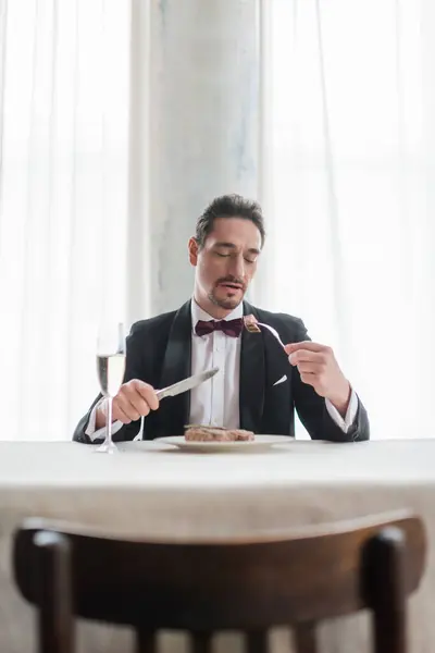 Gentiluomo ben vestito in smoking mangiare deliziosa bistecca di manzo vicino a un bicchiere di champagne, vita ricca — Foto stock