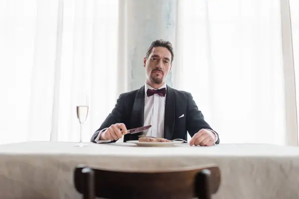 Rico caballero en esmoquin comiendo delicioso filete de res cerca de la copa de champán, mira a la cámara - foto de stock