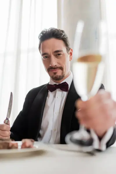 Hombre rico en esmoquin vitoreando copa de champán cerca de delicioso filete de carne de res y mirando a la cámara - foto de stock