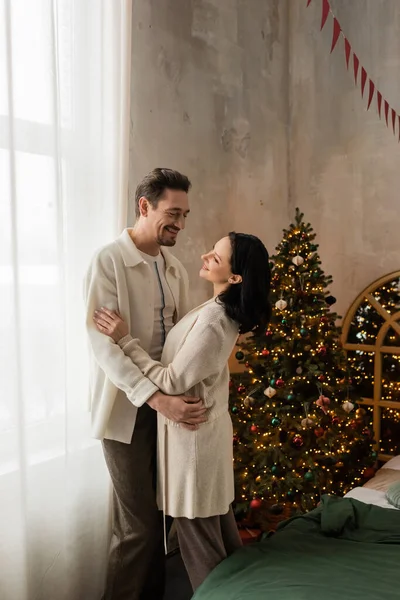Feliz pareja en casa cómoda llevar sonriendo y abrazando cerca del árbol de Navidad decorado en el dormitorio - foto de stock