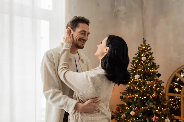 Freudiges Paar in bequemer Heimkleidung lächelt und umarmt neben geschmücktem Weihnachtsbaum im Schlafzimmer — Stockfoto