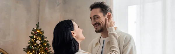 Alegre pareja en cómodo hogar llevar sonriendo y abrazando cerca decorado árbol de Navidad, pancarta - foto de stock