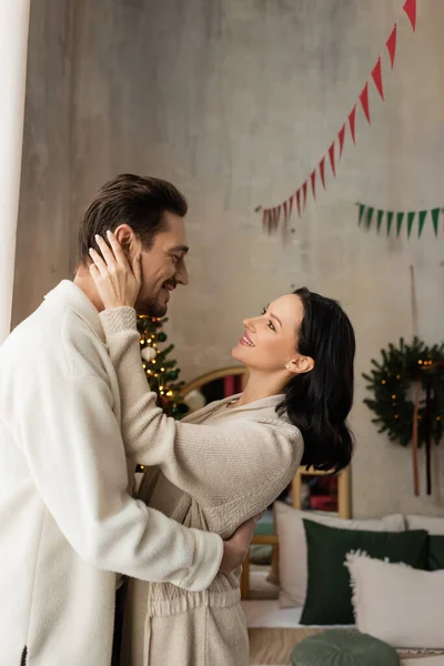 Fröhliche Frau in bequemer Hauskleidung umarmt Mann neben Weihnachtsbaum im Schlafzimmer, Freude — Stockfoto