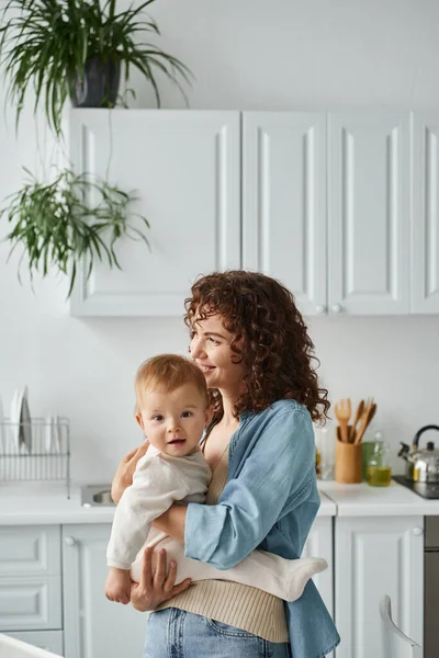 Donna gioiosa con i capelli ondulati abbracciando figlia bambino in cucina a casa, amore e tenerezza — Foto stock