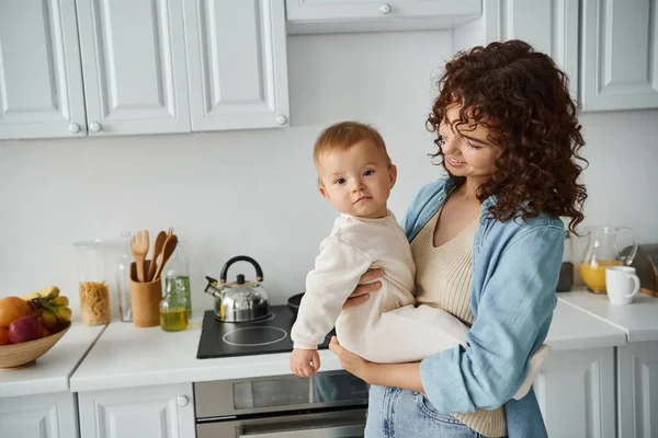 Smiling curly woman holding baby girl in romper looking at camera in cozy kitchen, happy family — Stock Photo